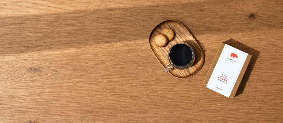 A kraft paper box with a white label sits on a light wood tabletop beside a small wooden tray holding a black coffee cup and two cookies. The box has a red elephant logo and the text 'The Elephant Coffee Co.