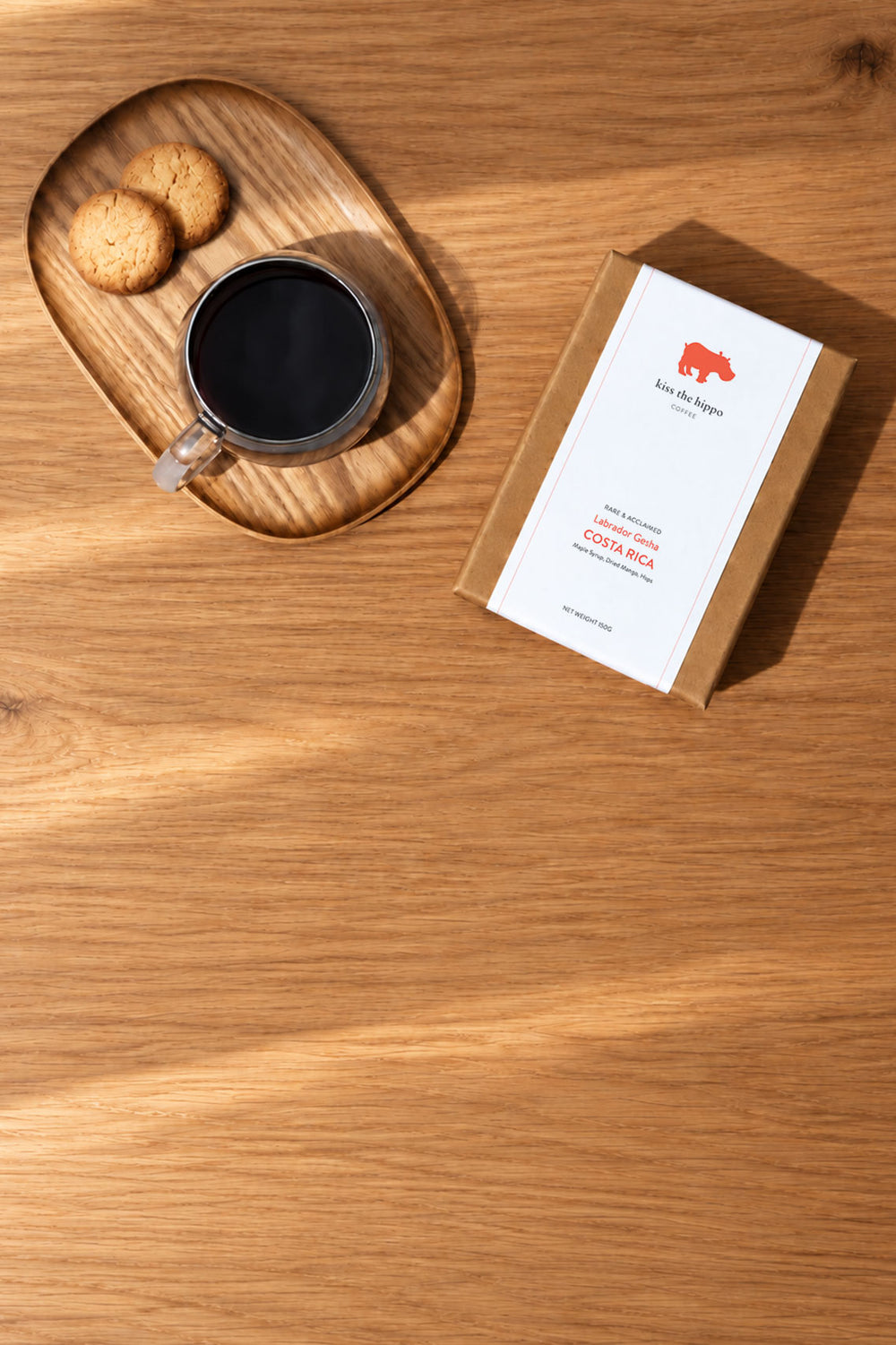 A kraft paper coffee box with a white label sits on a wooden table beside a cup of black coffee on a wooden tray and two cookies. The label shows a red rhinoceros logo and the text 'Kopi Daru'.