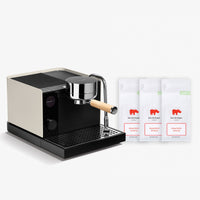 A modern espresso machine in cream and black with a stainless steel portafilter and wooden handle, shown next to three white coffee bean bags labeled 'Koe Die Koppie Roasted Coffee'.