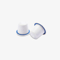 Two white coffee pods with blue rims, one standing upright and the other lying on its side, on a light gray background.