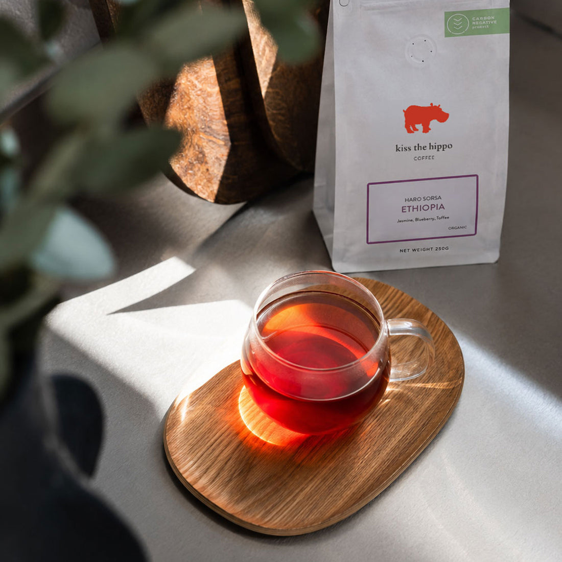 A bag of Kiss the Hippo Coffee Ethiopia whole bean coffee stands next to a clear glass cup filled with brewed red coffee on a wooden saucer, set in a softly lit environment with blurred green leaves in the foreground.