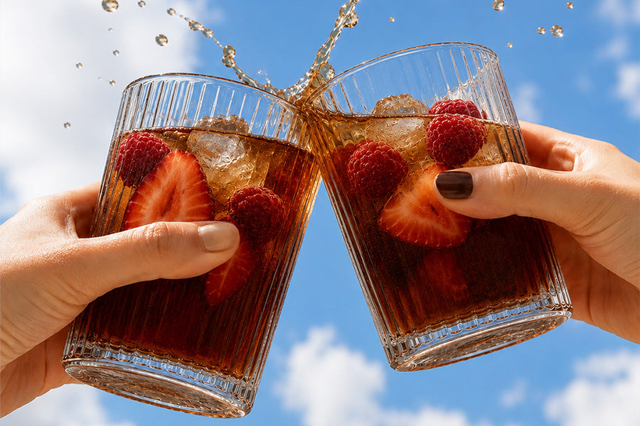 Two hands holding clear ribbed glass tumblers filled with a dark cola-like beverage, ice, strawberries, and raspberries, clinking together with a splash against a blue sky with clouds.