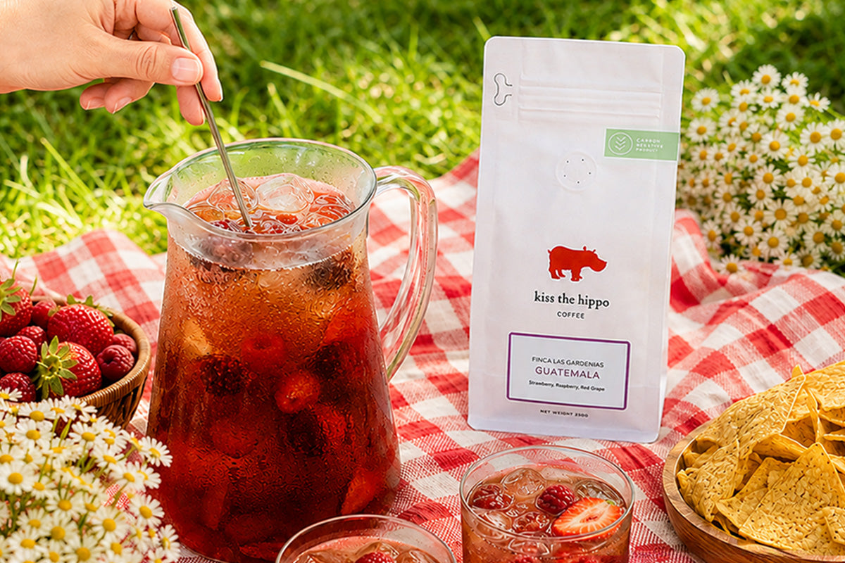 A white stand-up pouch of Kisu the Hippo Coffee sits on a red-and-white checkered blanket beside a large glass pitcher filled with iced strawberry tea, with strawberries, glasses, and flowers on grass in an outdoor picnic setting.